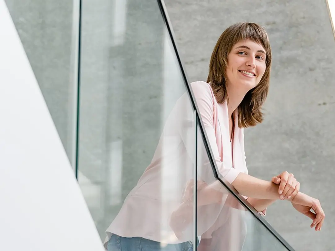 Frau in weißer Bluse und blauer Jeans lehnt an einem gläsernen Geländer in einem modernen, minimalistisch gestalteten Gebäude mit Betonwänden und viel Tageslicht.