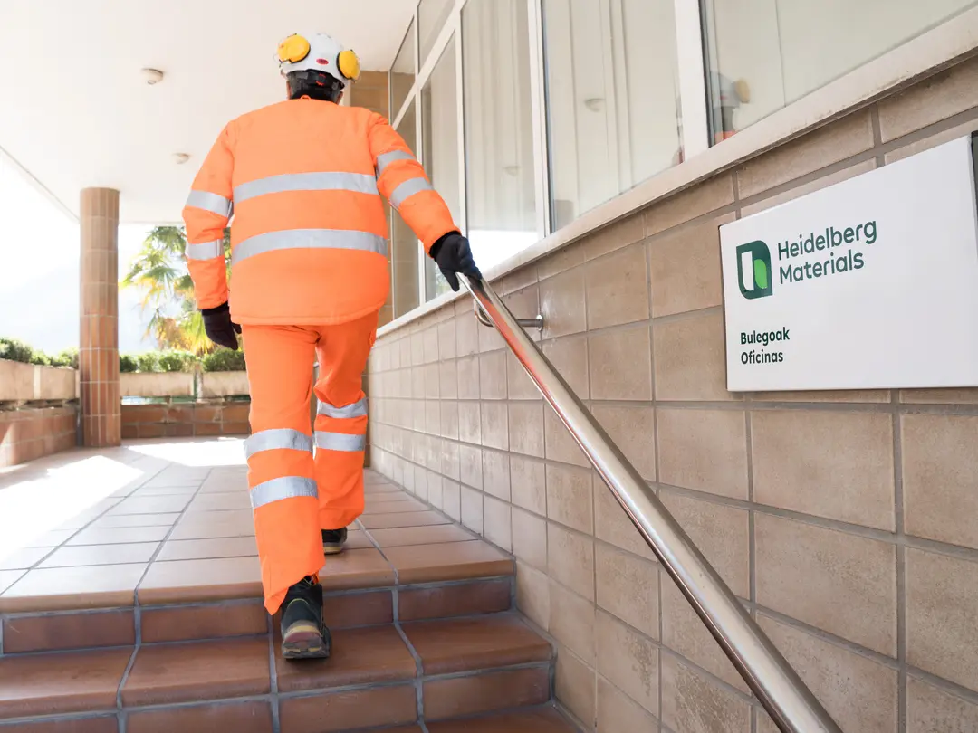 Eine Person in Schutzkleidung, von hinten zu sehen, geht eine Treppe hoch, an der Wand ist ein Schild von Heidelberg Materials