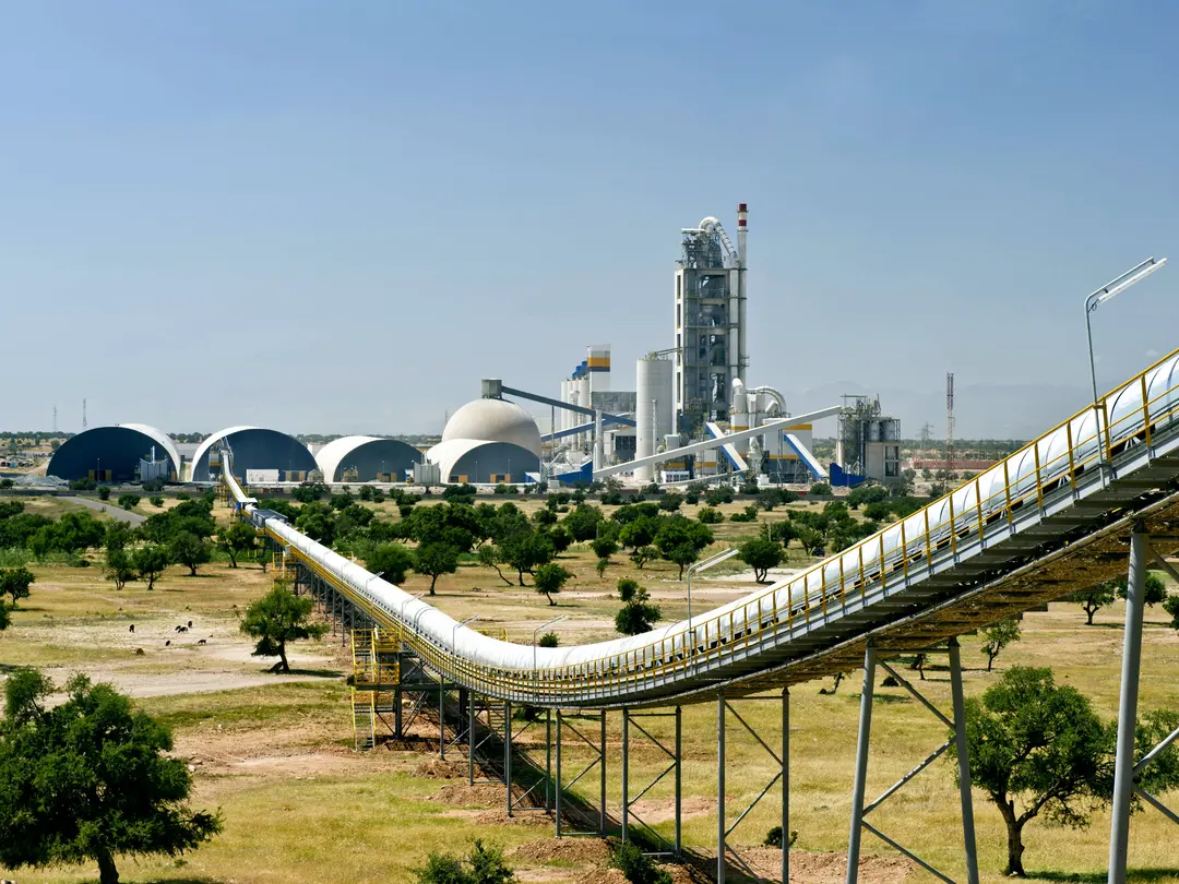 Industriegelände mit Zementfabrik und Förderband im Vordergrund, das diagonal von rechts unten nach links oben verläuft. Im Hintergrund sind mehrere Kuppelbauten, ein hoher Turm und Schornsteine sichtbar. Umgeben von einer trockenen Landschaft mit verstreuten Bäumen.