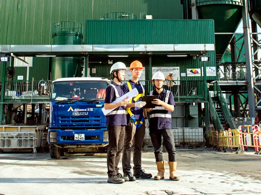Drei Arbeiter in Sicherheitsausrüstung, darunter Helme und Warnwesten, stehen auf einer Baustelle vor einer grünen Industrieanlage und einem blauen Lkw und prüfen Dokumente. In der Umgebung befinden sich Maschinen, Sicherheitsbarrieren und Baumaterialien.