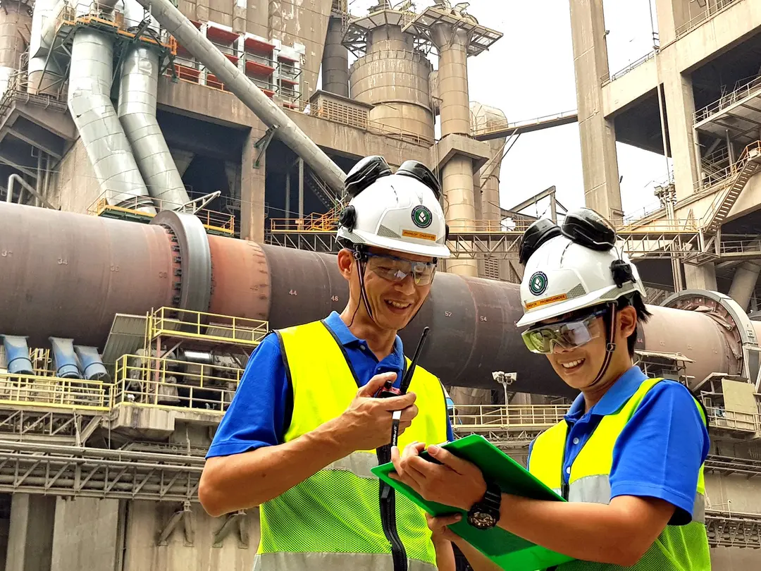 Zwei Industriearbeiter mit Schutzhelmen, Schutzbrillen und neonfarbenen Sicherheitswesten stehen im Vordergrund einer großen Industrieanlage mit Rohren und Maschinen. Einer hält ein Walkie-Talkie in der Hand, während der andere ein grünes Klemmbrett benutzt, was auf Kommunikation und Aufgabenkoordination hindeutet.