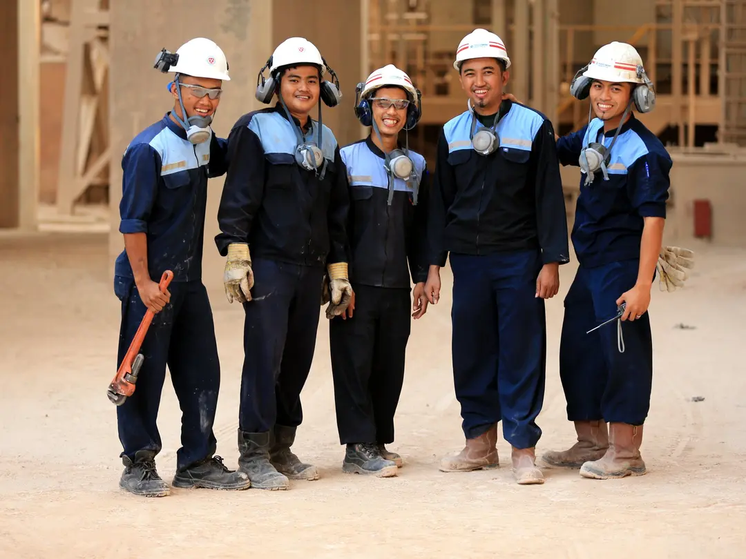 Eine Gruppe von fünf Bauarbeiter*innen in dunkelblauer Arbeitskleidung und weißen Helmen steht in einem Rohbau. Sie tragen Schutzbrillen und Atemschutzmasken, einige halten Werkzeuge wie einen Schraubenschlüssel und eine Zange.