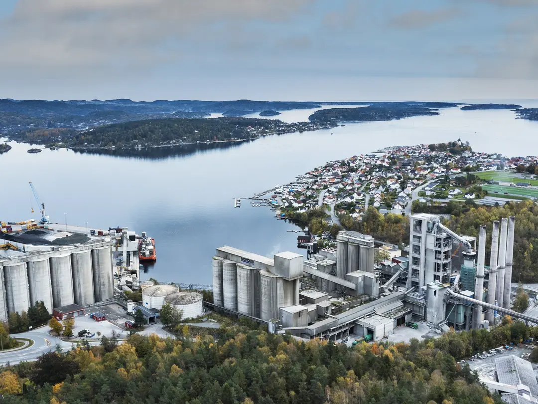 Luftaufnahme des Zementwerks Brevik am Meer, umgeben von Wäldern und einer Kleinstadt. Links sind Silos und Hafenkräne sichtbar, rechts Industriegebäude mit Schornsteinen.