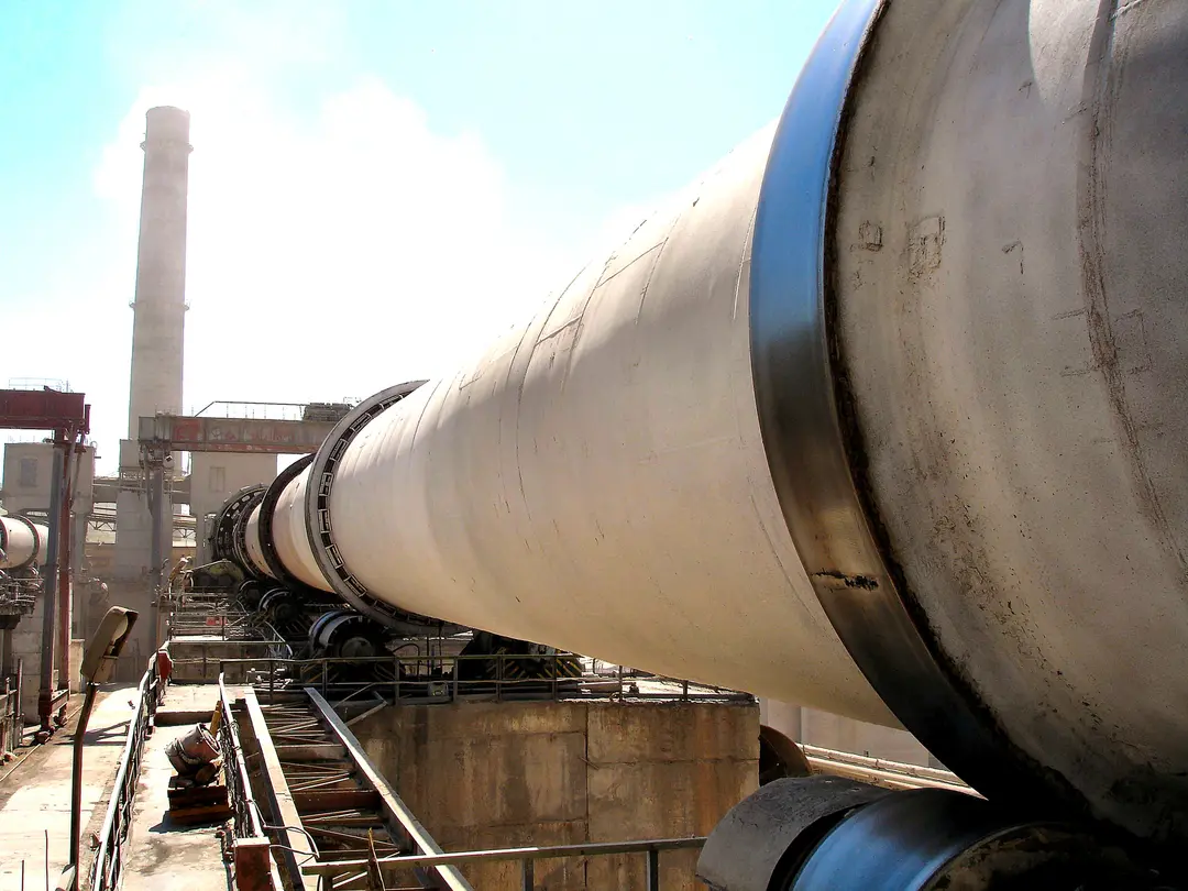 Drehrohrofen, lange graue, leicht geneigte Röhre, die sich perspektivisch nach hinten verjüngt, dahinter ein Schornstein vor einem hellblauen Himmel