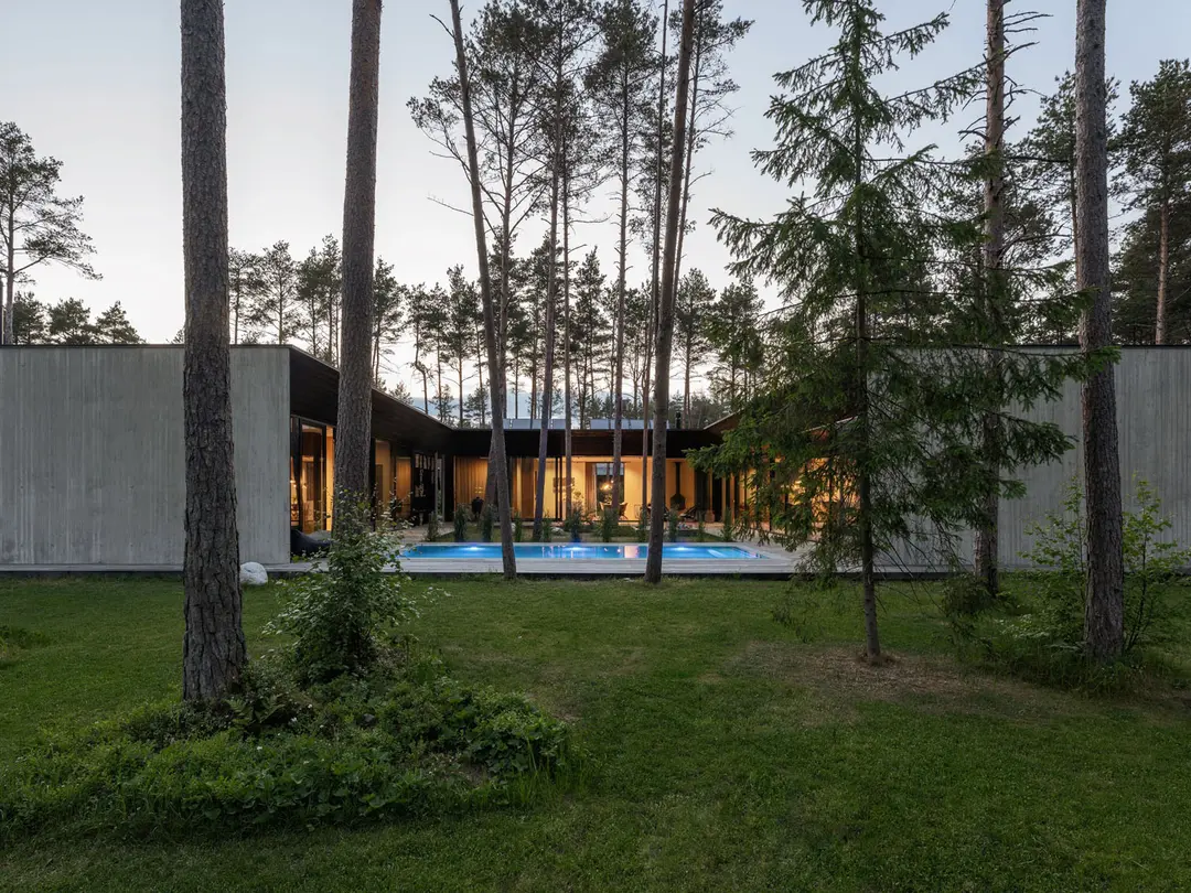 Bungalow aus Sichtbeton, umgeben von einzelnen Bäumen, im Innenhof ist ein beleuchteter Pool zu sehen