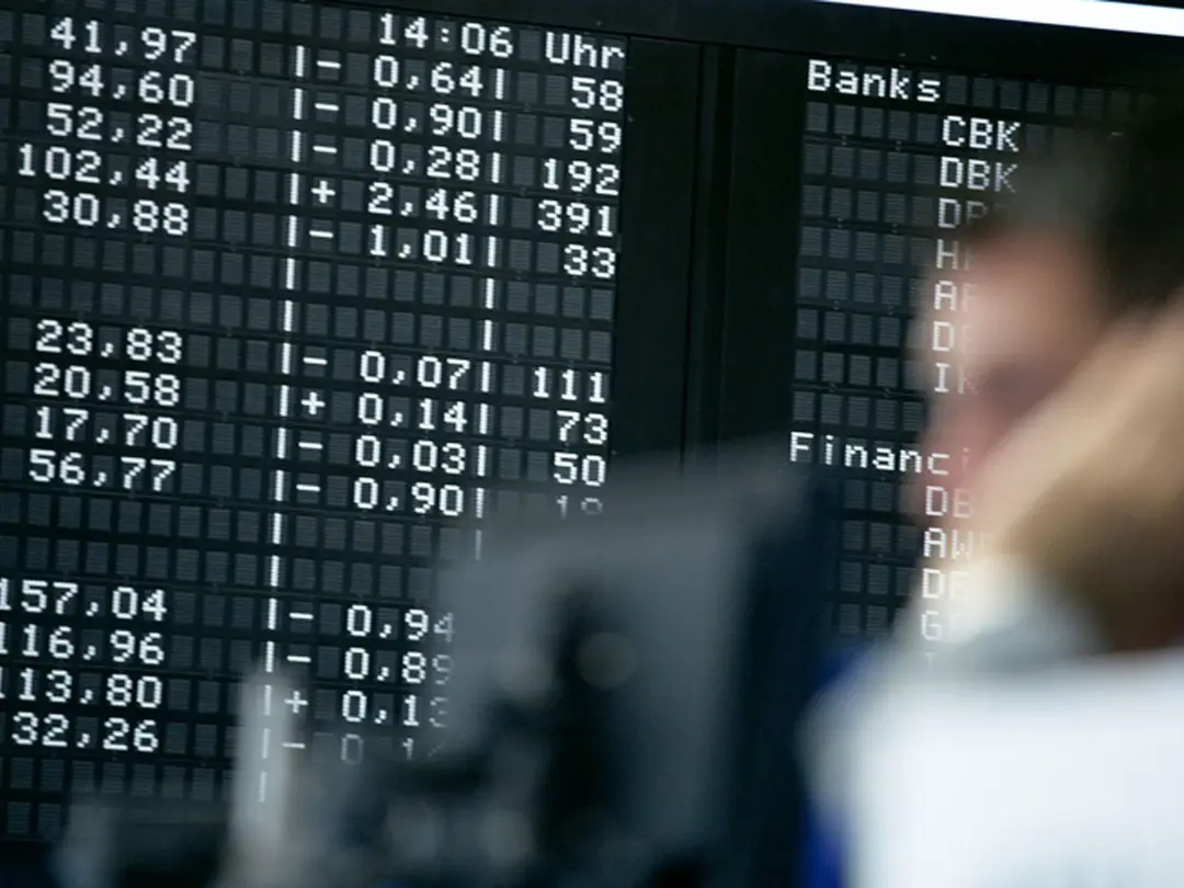 Eine verschwommene Figur im Vordergrund mit einer digitalen Börsentafel im Hintergrund, die verschiedene Zahlen und Aktiencodes anzeigt.