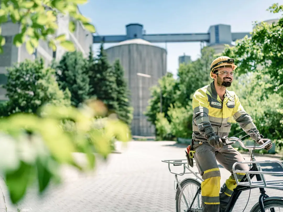 A person in yellow and black work attire with a helmet rides a bicycle. Large industrial silos and lush green trees are visible under a clear blue sky in the background.