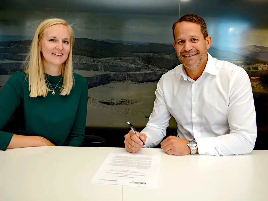 A woman in a green dress and a man in a white shirt are sitting at a table, a piece of paper between them, the man is holding a pen in his hand.
