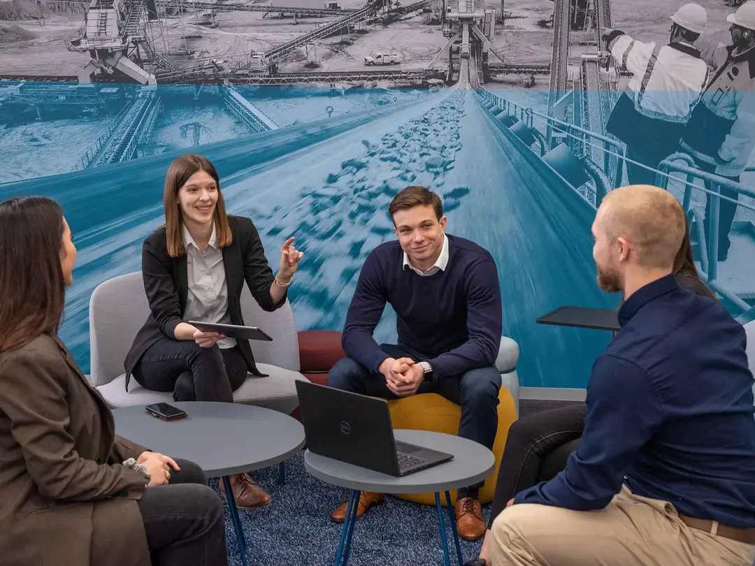 Vier junge Menschen im Business-Casual-Look sitzen um einen niedrigen runden Tisch herum, im Hintergrund das Bild eines blau eingefärbten Förderbandes