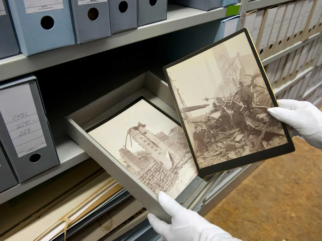 Hände mit Handschuhen halten eine Schachtel mit historischen Fotos, eine Hand hält dabei eines der Fotos