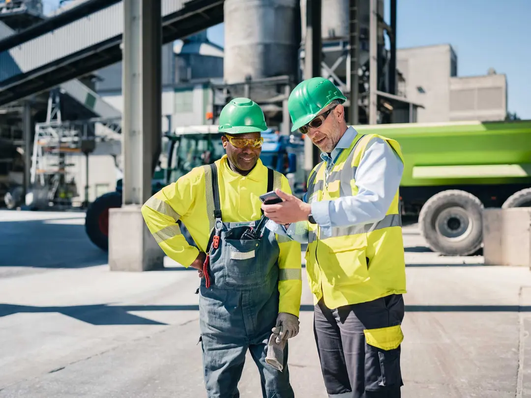 Zwei Männer mit Helmen stehen vor einem Zementwerk, eine Person zeigt der anderen etwas auf einem Smartphone.
