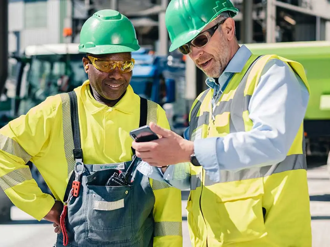 A construction worker in a yellow vest shows their colleague something on his mobile phone