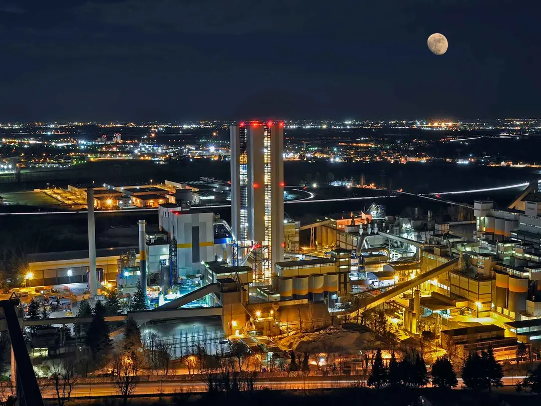 Zementwerk bei Nacht mit beleuchteten Anlagen und hohen Silotürmen, umgeben von städtischer Landschaft und einem Vollmond im Hintergrund.