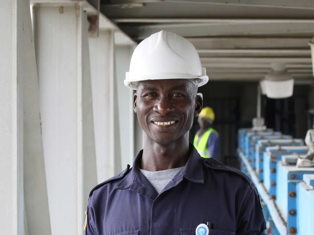 Arbeiter in blauem Hemd mit weißem Helm auf einer Baustelle, im Hintergrund eine weitere Person