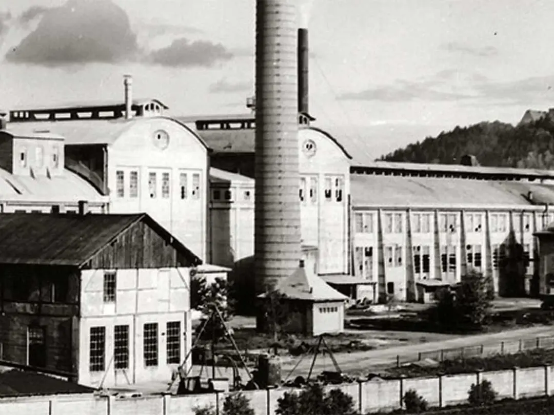 Historisches Schwarz-Weiß-Foto eines Industriekomplexes mit markantem Schornstein vor Hügelkulisse