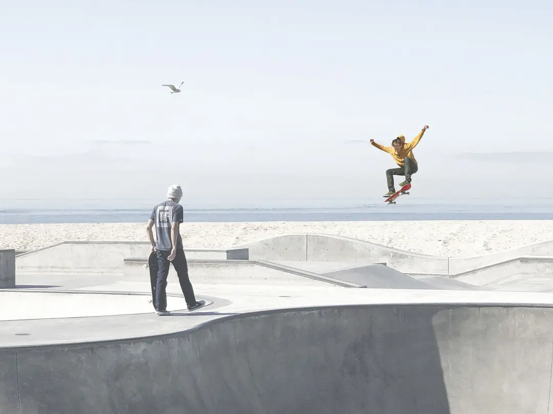 Skater springt mit Skateboard in einer leeren Skatepark-Bowl, während eine andere Person am Rand steht.