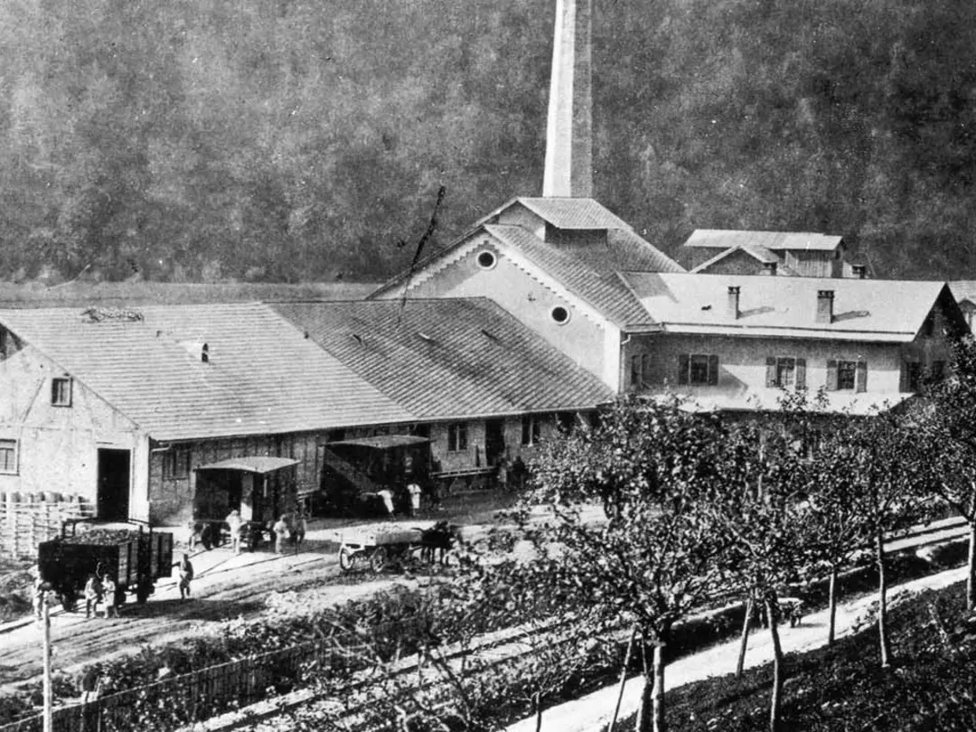 Historisches Foto industrieller Hallen mit Schloten, davor Güter-Waggons, Kutschen und Menschen