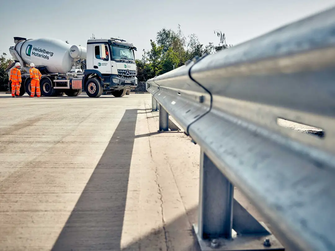 A ready-mixed concrete truck is parked on a paved surface, with two workers in safety vests standing nearby. A metal guardrail is visible in the foreground.