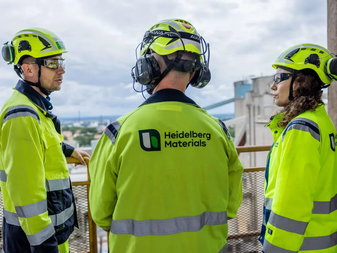 Drei Personen in Schutzkleidung stehen an einem Geländer auf einer Plattform eines Zementwerks, im Hintergrund eine wolkenverhangene Landschaft