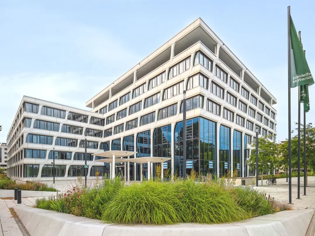 White office building, greenery in front, three flagpoles with green flags next to it