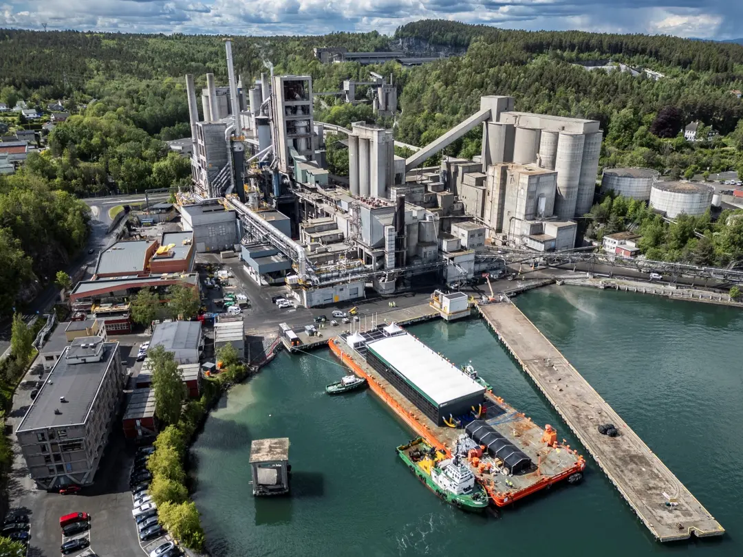Aerial view of a cement plant with silos, conveyors, and processing units, located near a port with a docked barge for material transport.