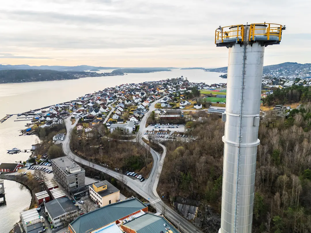 Luftaufnahme eines hohen Industrieschornsteins mit einem gelben Sicherheitsgeländer an der Spitze rechts. Unten verbindet eine kurvenreiche Straße ein Industriegebiet mit einer Wohnstadt mit dichter Bebauung. Die Stadt liegt in der Nähe eines ruhigen Ufers und ist von Waldgebieten umgeben, mit Hügeln im Hintergrund unter einem bewölkten Himmel.