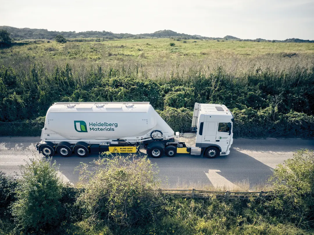 Ein weißer LKW mit Auflieger und dem Logo "Heidelberg Materials" fährt auf einer schmalen Landstraße, umgeben von grüner Vegetation und einer weitläufigen Landschaft im Hintergrund.