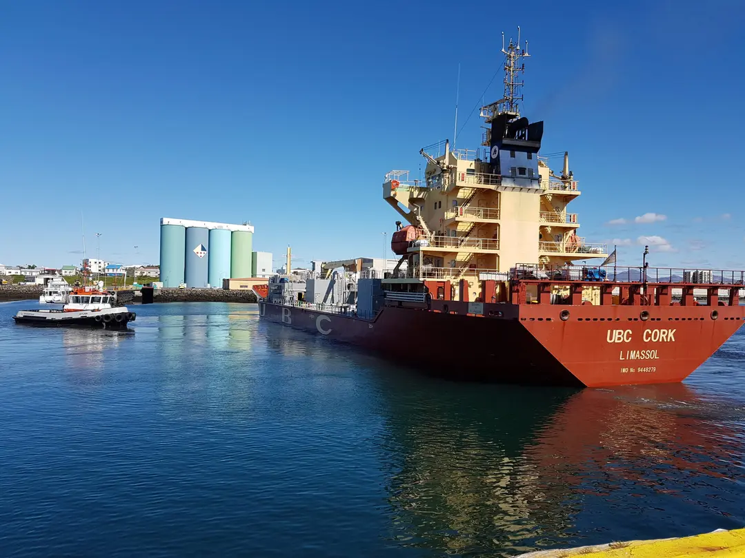 Ein rotes Frachtschiff fährt im Hafen ein, begleitet von einem kleinen Schlepperboot auf der linken Seite. Im Hintergrund sind ein Lagergebäude und der blaue Himmel zu sehen. Im Vordergrund eine gelb gestrichene Kaimauer.