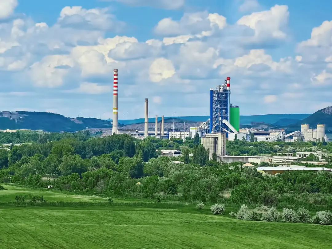 Panorama einer grünen Landschaft mit dem Zementwerk von Devnya in der Bildmitte; die Anlage umfasst mehrere Schornsteine und ein großes, blau-grünes Gebäude. Im Vordergrund weite Wiesen und Bäume, im Hintergrund Hügel und ein bewölkter blauer Himmel.