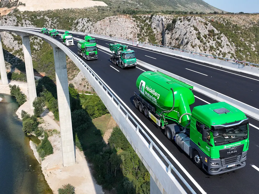 Grüne Betonmischer-Lkws fahren auf einer modernen Brücke, die über eine Schlucht mit Fluss führt. Im Hintergrund sind Berge und Natur sichtbar. Die Fahrzeuge sind in einer Kolonne angeordnet und zeigen ein firmenspezifisches Design.