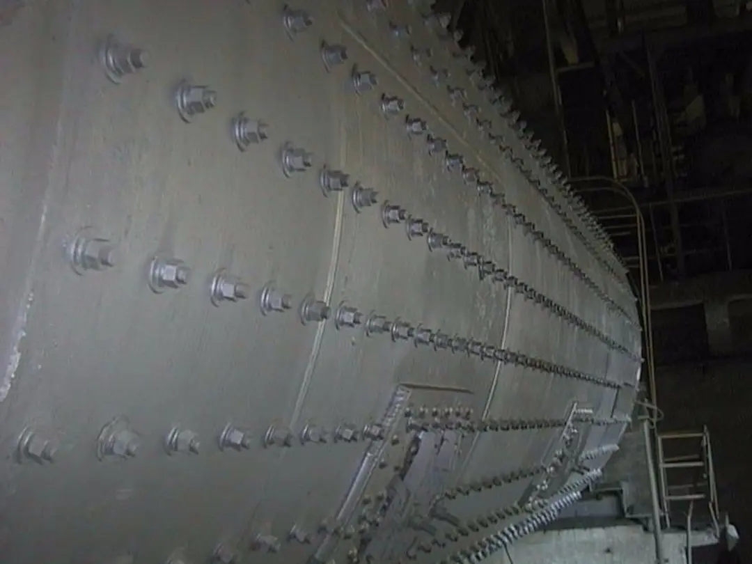 Nahaufnahme einer Zementmühle mit Reihen gleichmäßig laufender Bolzen, die diagonal von links unten nach rechts oben angeordnet sind. Im Hintergrund rechts sind eine Metallleiter und ein Gerüst zu sehen.