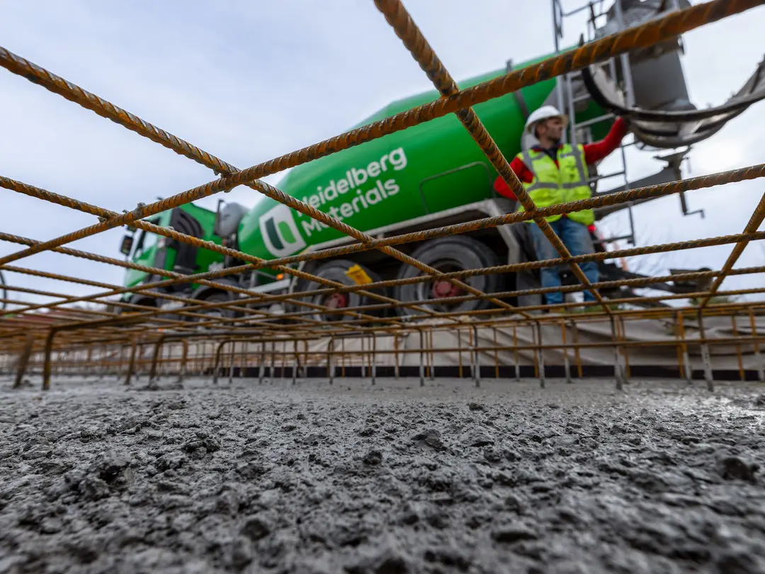 Blick auf eine Baustelle: Im Vordergrund frisch gegossener Beton, durchzogen von Stahlarmierungen. Im Hintergrund ein grüner Betonmischer von Heidelberg Materials, daneben ein Bauarbeiter mit Sicherheitsweste und Helm, der eine Betonrutsche bedient.