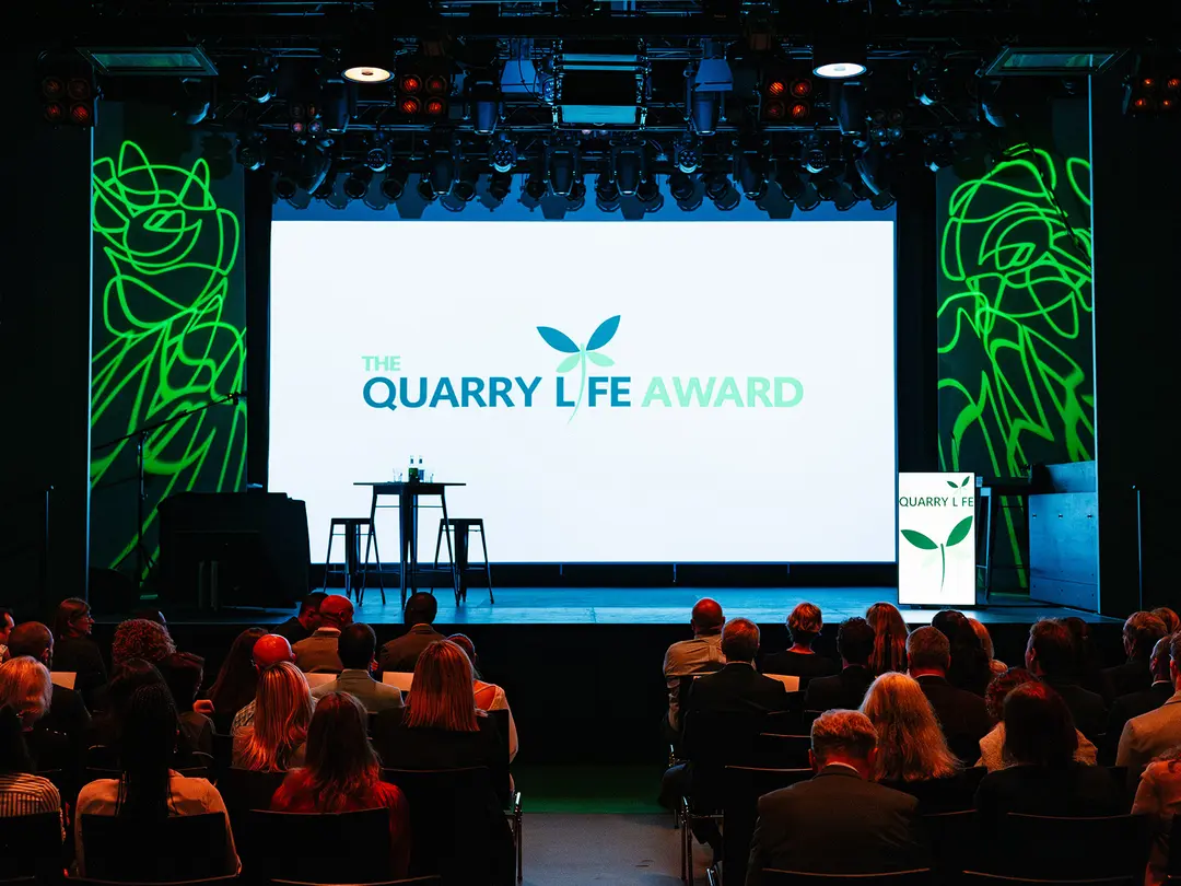 Bühne mit großer Leinwand, auf der "The Quarry Life Award" steht; davor zwei leere schwarze Rednerpulte und ein Rednerpult rechts mit Logo; Publikum sitzt in mehreren Reihen, Hintergrund mit grünen, beleuchteten Linienmustern.
