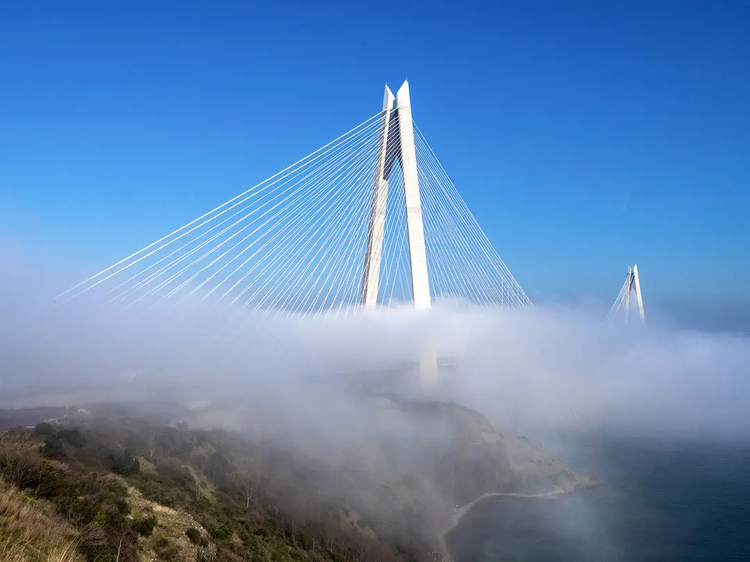 Cable-stayed bridge emerging from fog over a coastal landscape, showcasing modern infrastructure.