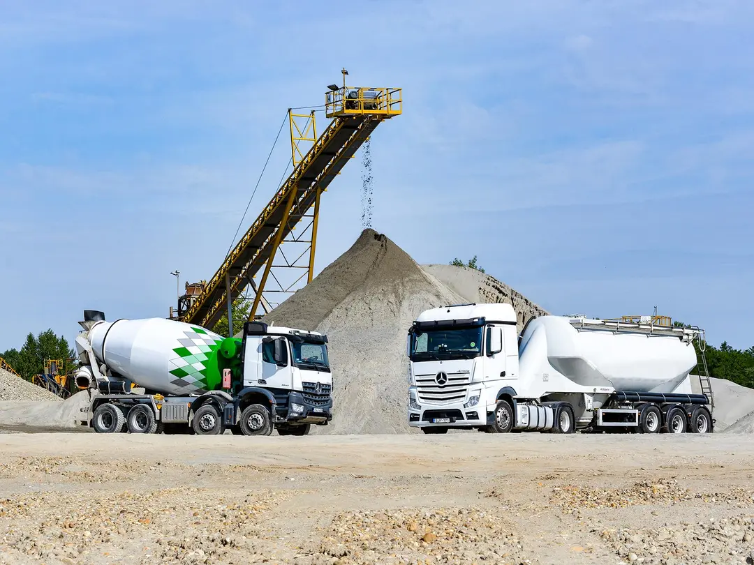 Ready-mixed truck and silo truck standing in front of a sand heap, sand trickles down from a conveyor belt