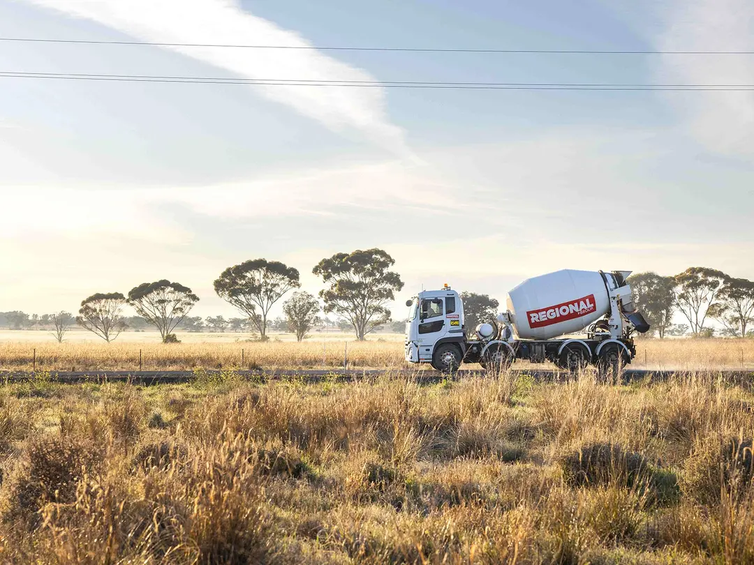 A ready-mixed concrete truck drives through a rural landscape with tall dry grass in the foreground and scattered trees under a lightly clouded sky in the background.