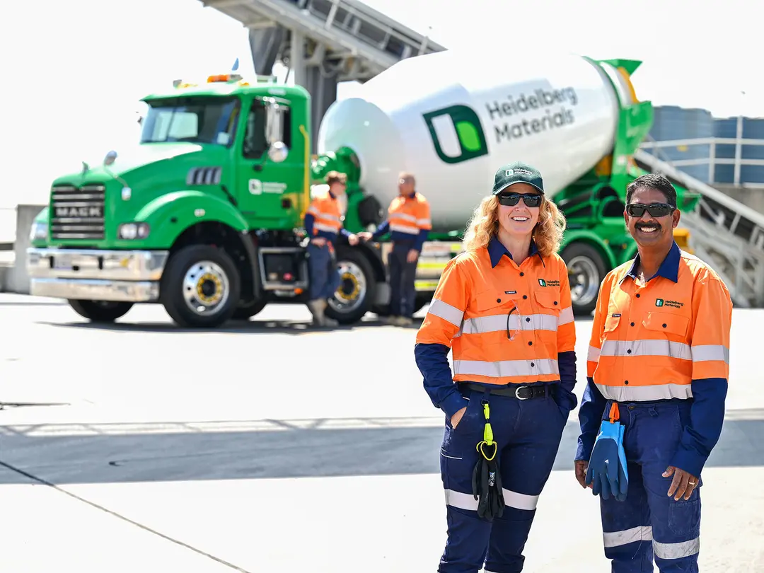 Two workers wearing orange safety vests and hard hats stand in the foreground, smiling. Behind them a green and white Heidelberg Materials RMC truck.