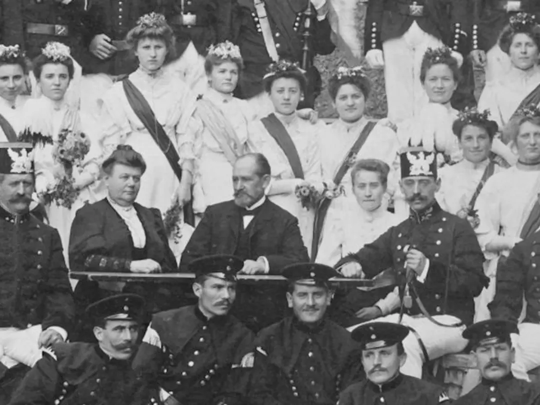 Historical photo with darkly dressed men and lightly dressed women standing in alternating rows behind each other.
