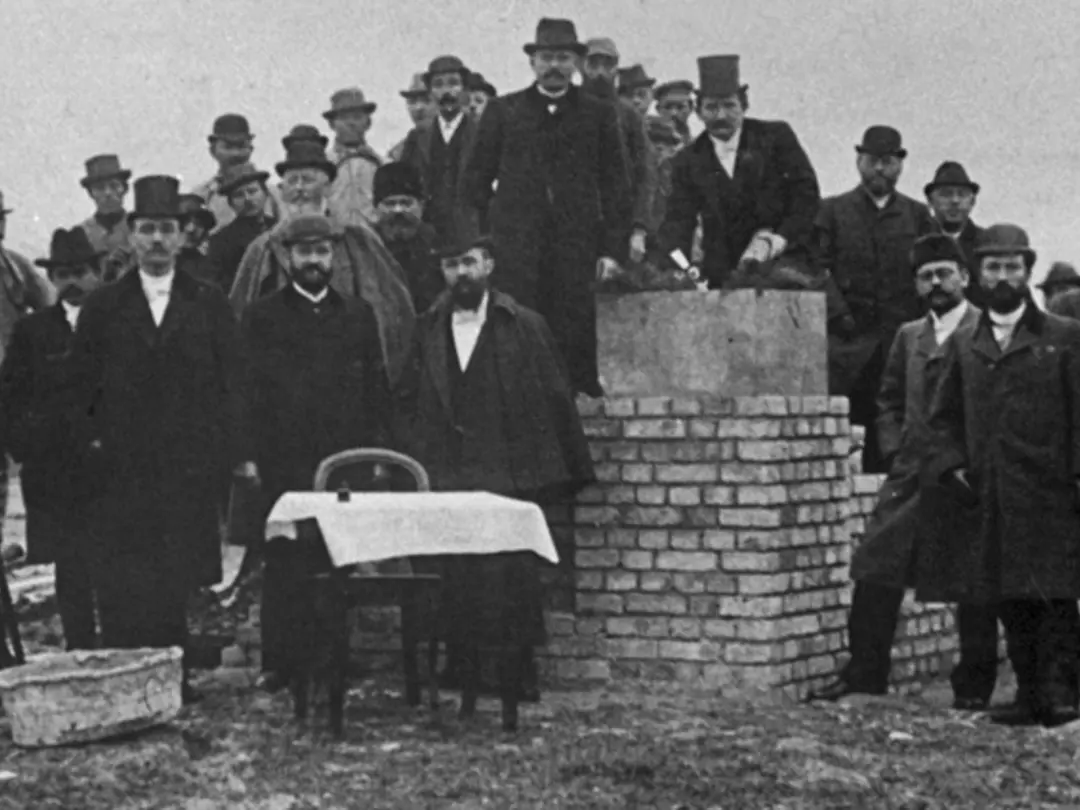 Großgruppe von Menschen bei einer Grundsteinlegung auf einer Baustelle im historischen Kontext.