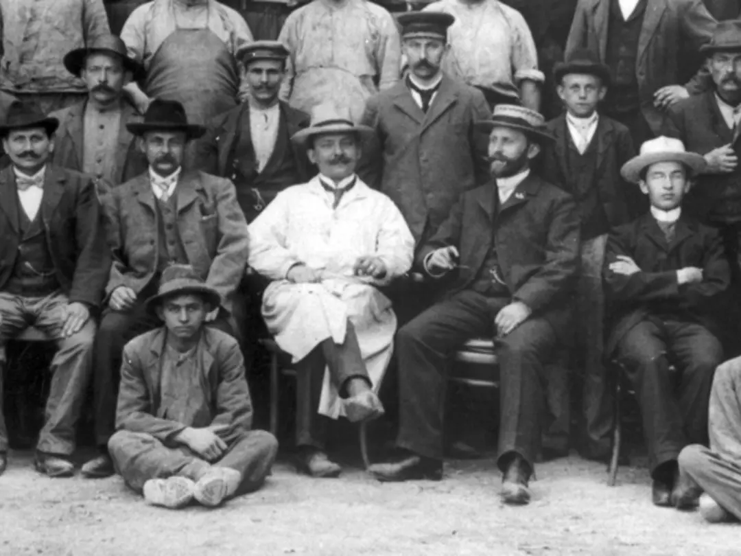 Historische Gruppenaufnahme von Menschen vor einem Gebäude, umgeben von Fässern mit Firmenlogo.