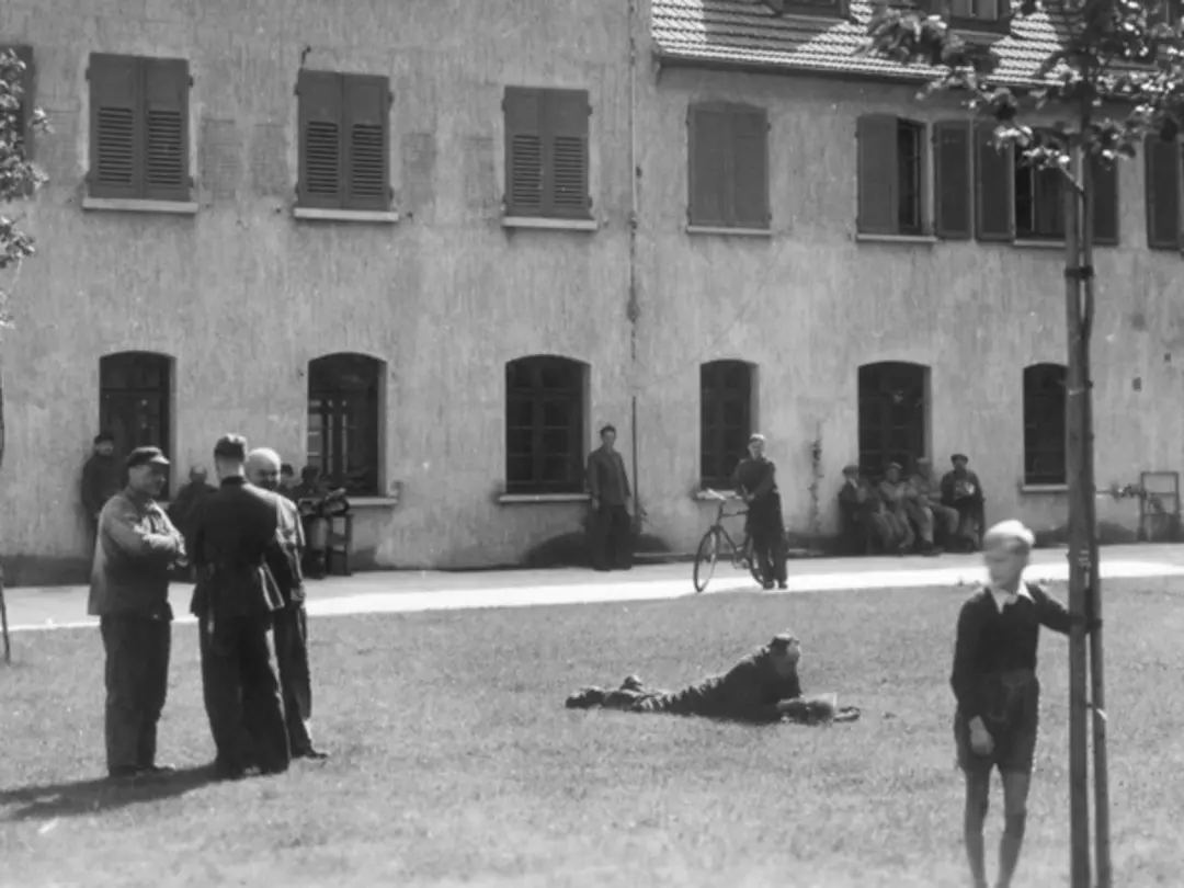Historische Szene mit Menschen auf einem Platz vor einem Gebäude, im Hintergrund ein Wasserbrunnen.