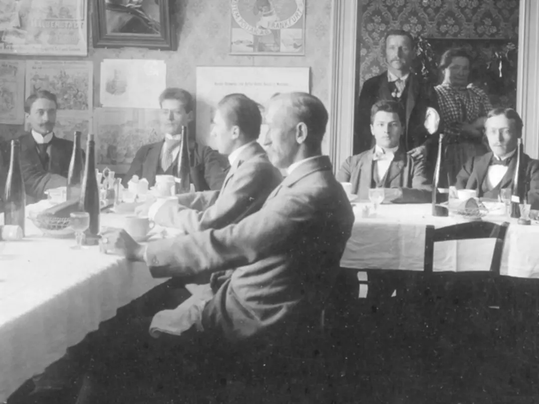 Historisches Foto von Männern in Anzügen, die um einen Tisch sitzen, auf dem Weinflaschen stehen. Im Hintergrund steht eine Frau im Türrahmen.