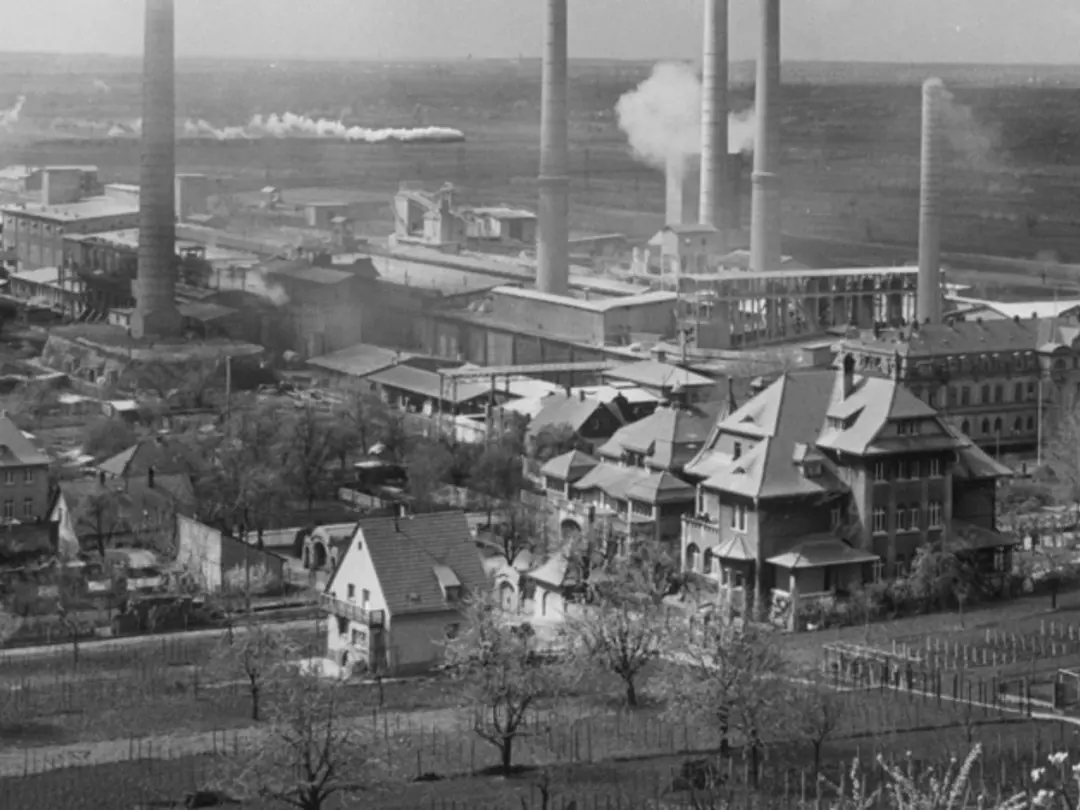 Historische Industrieanlage mit mehreren Schornsteinen und umliegenden Gebäuden in einer ländlichen Umgebung.