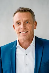 Christoph Beumelburg wearing a white shirt and a blue jacket, in the background a window and an exposed concrete wall