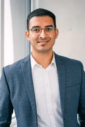 Samuel Jacob in weißem Hemd und grauem Jackett vor einer Sichtbetonwand und einer Fensterfront