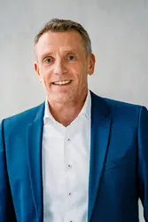 Christoph Beumelburg mit weißem Hemd und blauem Jackett, im Hintergrund ein Fenster und eine Sichtbetonwand