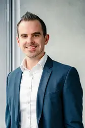 Steven Meyers in dunkelblauem Anzug und weißen Hemd vor einer Sichtbetonwand, neben einem Fenster