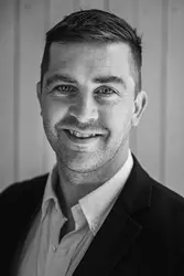 A man wearing a dark blazer and light dress shirt, standing against a vertical wooden panel background.