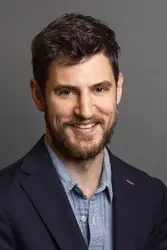 A person wearing a blazer and shirt is posed against a plain background.