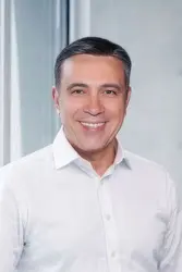 Ozan Kacar wearing a white shirt standing in front of a window, one hand on the windowsill